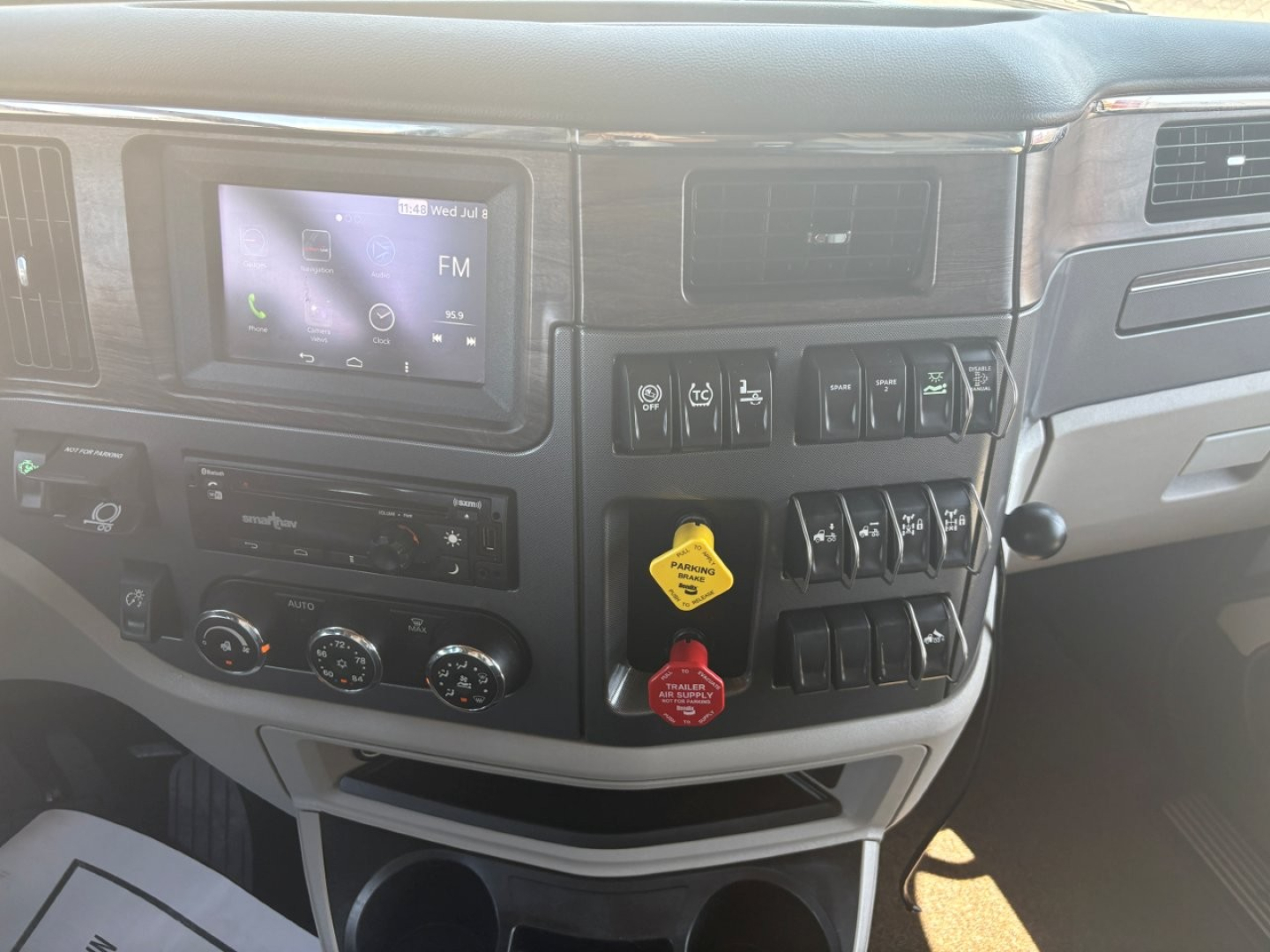 Peterbilt 579 truck dashboard interior showing infotainment screen, climate controls, and multiple toggle switches; center lower dash features two circular knobs and a yellow parking brake handle with red tag.