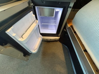 Truck cab interior showing a compact refrigerator/freezer unit with open door and empty shelves, located between driver seat and passenger seat area.
