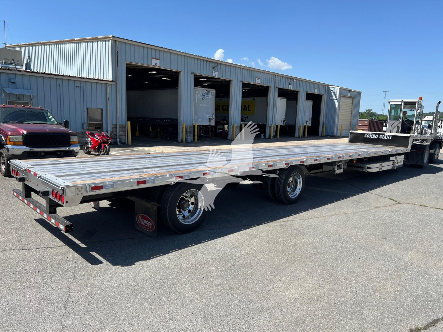 Flatbed trailer with ramp kit attached, long aluminum deck, two-axle configuration, shown in a parking lot with a warehouse in the background.
