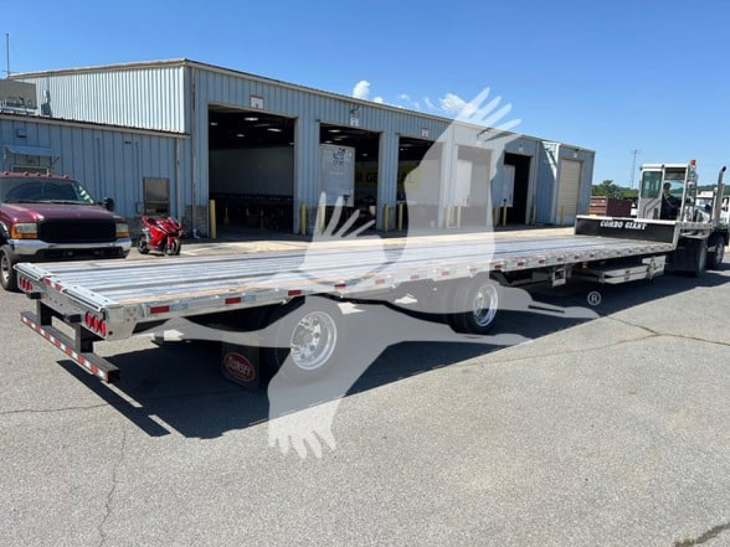 Flatbed trailer with ramp kit and long drop deck showing metal deck, two axles, and hitch at rear. In a dealership lot with blue sky.