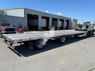 Flatbed trailer with ramp kit attached, long aluminum deck, two-axle configuration, shown in a parking lot with a warehouse in the background.