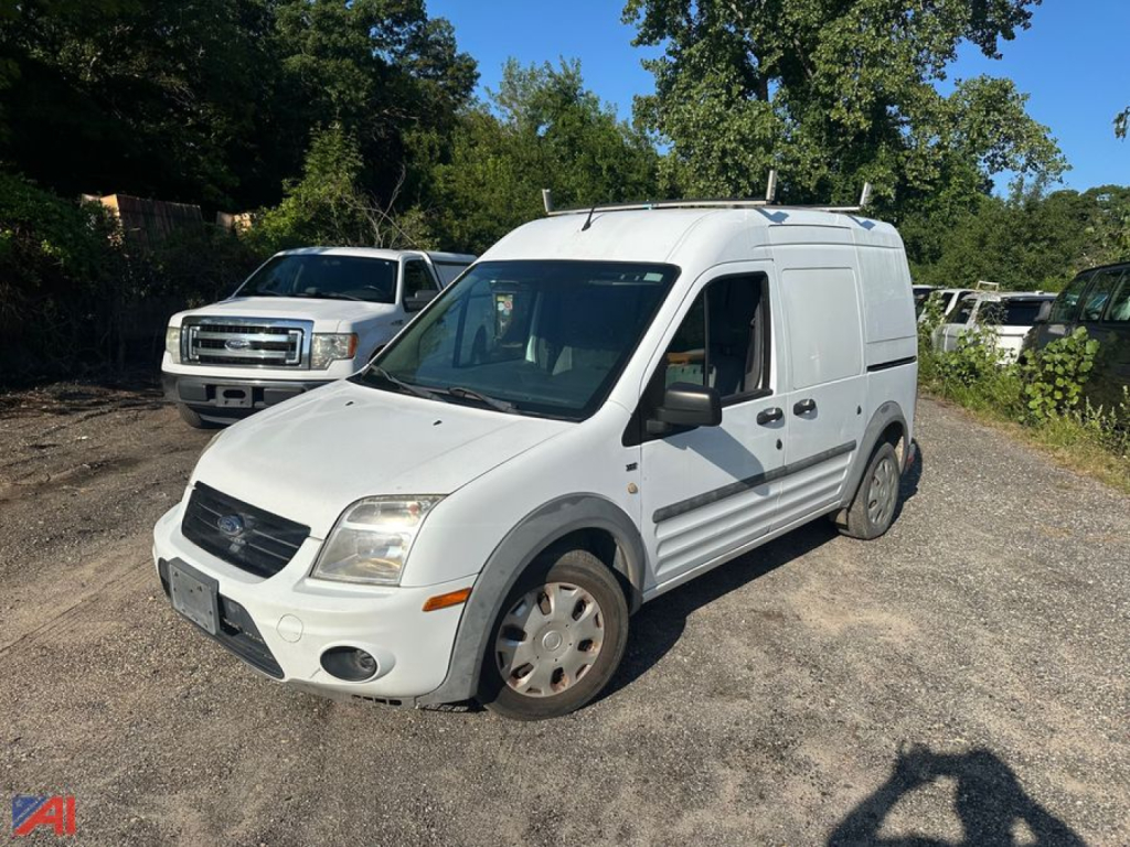 Ford Transit Connect cargo van in a parking lot viewing three-quarters front; white body, front grille, headlights, side door, roof rack with antennas; wheel hubs and gravel surface.