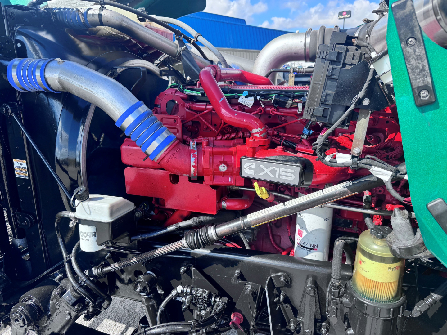 Kenworth W900L engine bay showing X15 logo on red engine, blue silicone hoses, white filters, and surrounding components.