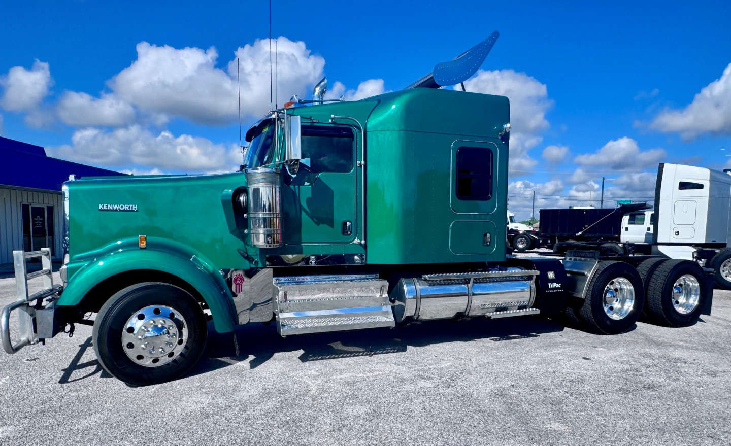 Green Kenworth W900L conventional sleeper truck side view showing chrome fuel tanks and dual rear axles under a blue sky.