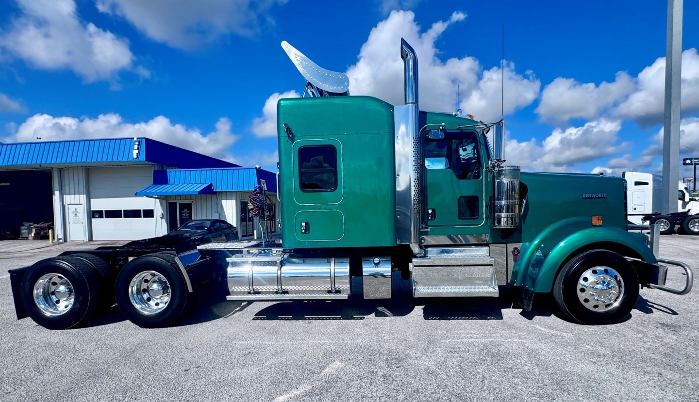 Kenworth W900L truck side view showing green cab, chrome fuel tanks, dual rear axles, and aerodynamic exhaust stacks.
