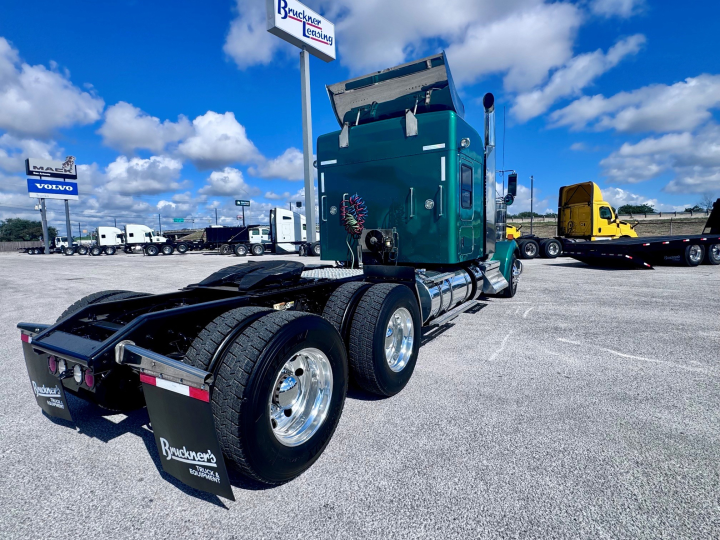 Kenworth W900L tractor in a parking lot, side view of rear wheels and green cab, dual rear axle setup, mud flaps with dealer branding, blue sky with clouds.
