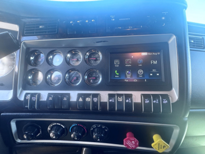 Kenworth W900L climate and gauge cluster with aftermarket touchscreen head unit displaying FM, and various switches below; dashboard and instrument panel visible.