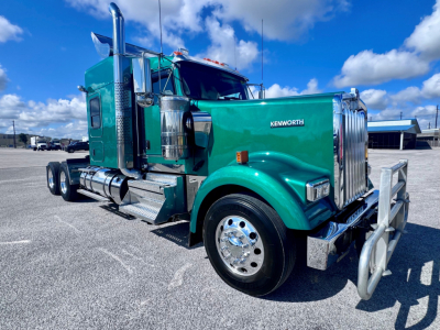 Kenworth W900L truck in green color, front-right view showing chrome bumper, large fuel tanks, and dual rear axles.
