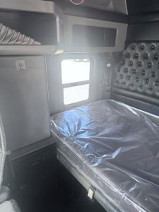 Kenworth W900L sleeper cab interior showing bed with plastic cover and quilted black headboard, side window, overhead storage compartments, and small controls.