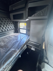 Kenworth W900L sleeper cab interior with window and bed visible; plastic wrap on mattress; side panels and storage compartments.