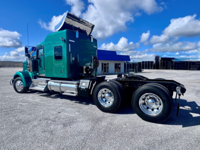 Kenworth W900L semi-truck in teal cab with sleeper, view of side showing dual rear wheels and chassis, chrome fuel tanks, and raised hood area.