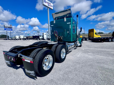Kenworth W900L tractor in a parking lot, side view of rear wheels and green cab, dual rear axle setup, mud flaps with dealer branding, blue sky with clouds.