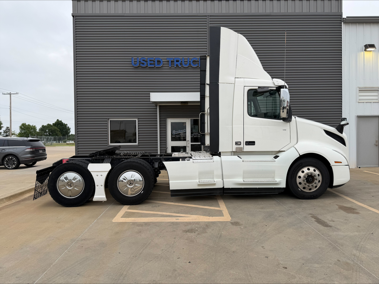 2019 Volvo VNL300 conventional daycab tractor truck side view showing sleeper cab and dual rear axles in front of a used truck dealership building.