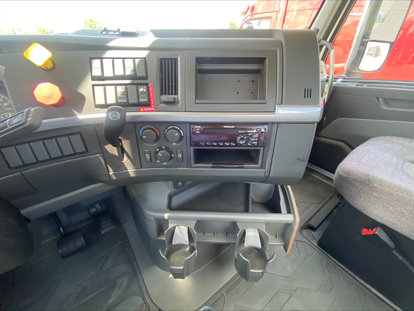 Volvo VNL cockpit dash showing steering column, gear shifter, center console with controls, cup holders, and dashboard vents in a daycab truck.