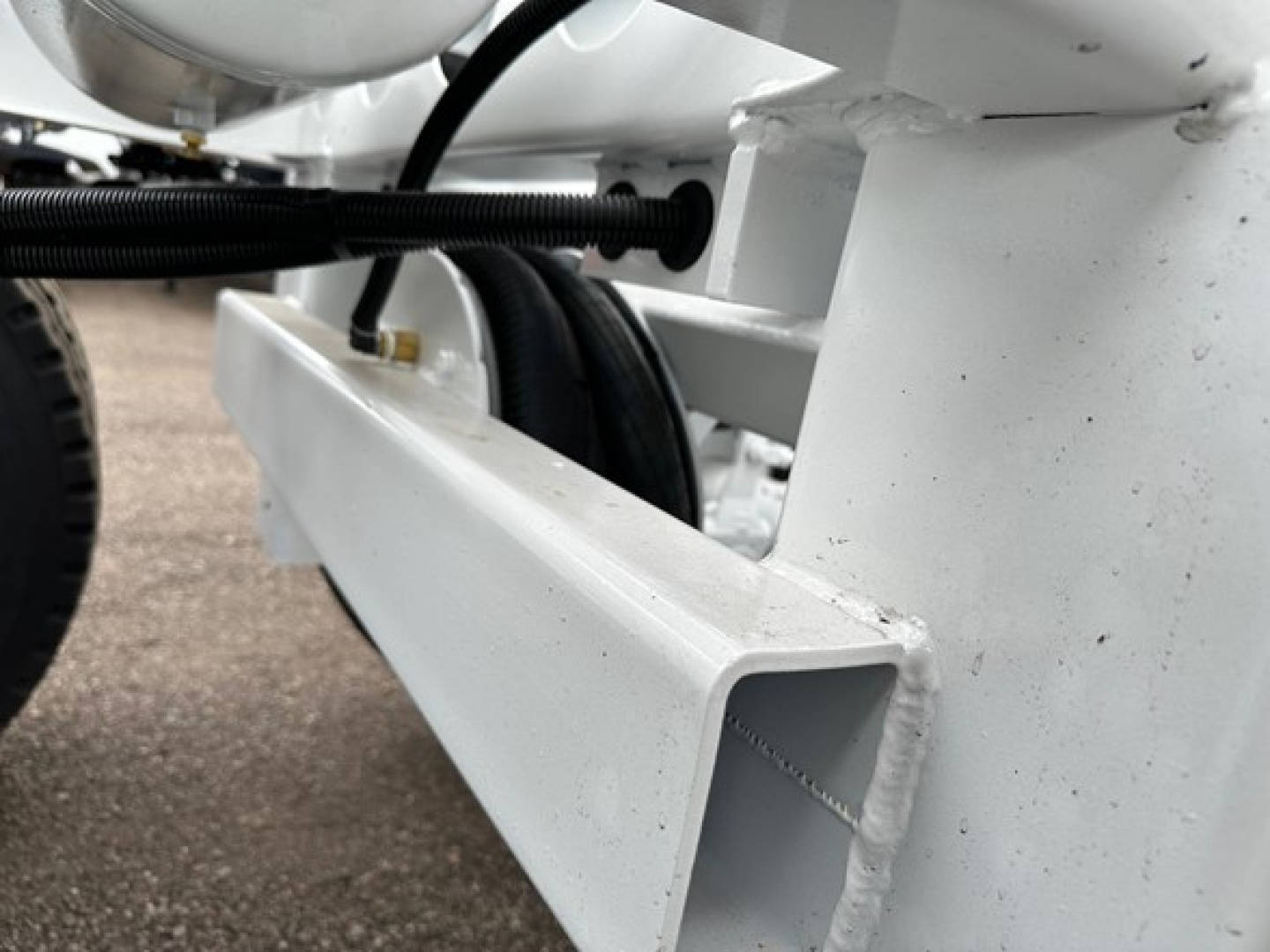 White trailer frame with structural I-beam and hydraulic hose assembly showing a black corrugated hose and a metal bracket on the trailer chassis.