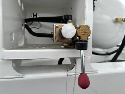 Valve assembly with brass block and lever on white trailer chassis, hydraulic line connected, rounded red protective cap on lever. Visible components: brass valve with markings, silver knob, black hose, metal rod and red rubber handle.