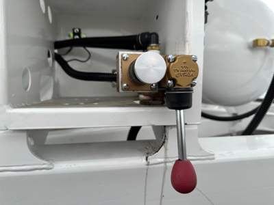 Dump trailer component close-up showing brass valve with label and red-knob grease fitting, mounted in white trailer frame, hydraulic line and fittings visible.