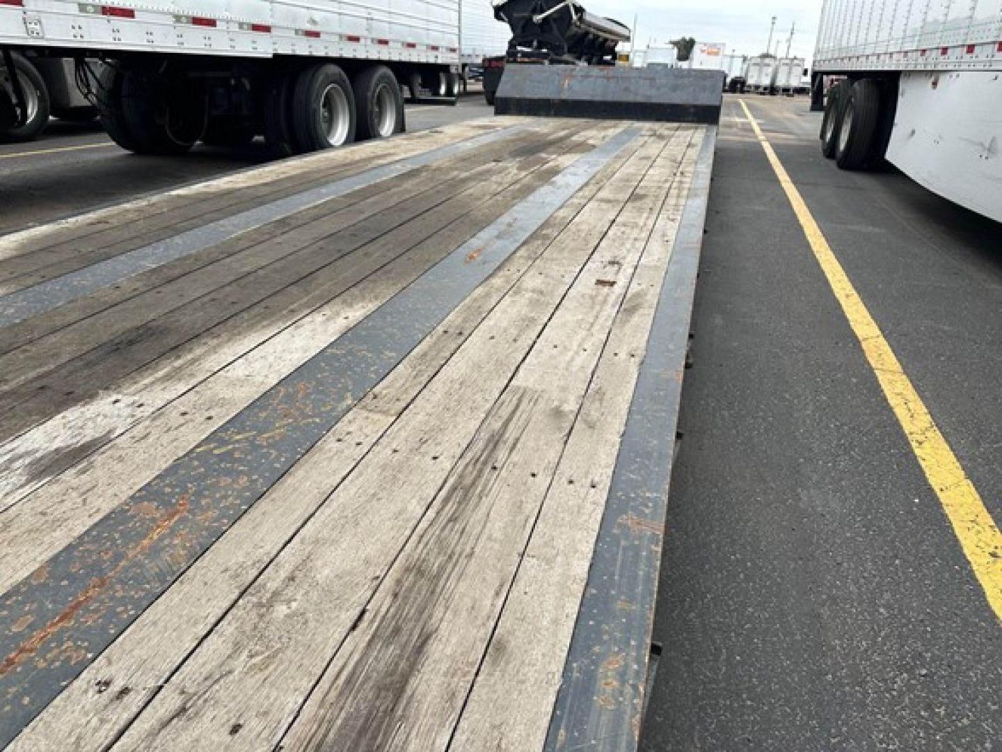 Flatbed trailer bed with wooden planks and metal side rails, viewed from the rear with other trailers parked on both sides.