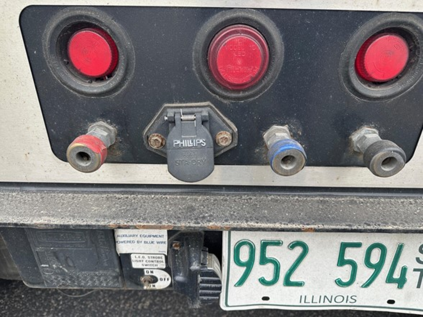 Trailer rear showing hydraulic connections and electrical sockets with red circular lights; includes 5th wheel or hitch area and Illinois license plate 952 5949.