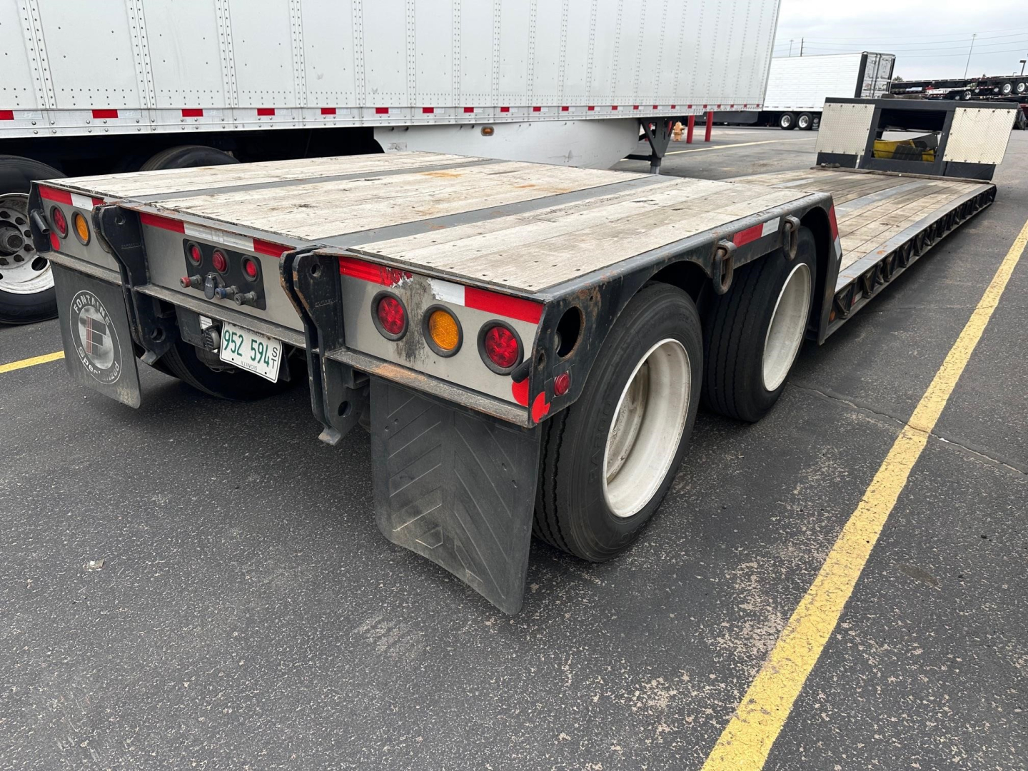 Flatbed trailer rear view showing triple axle with mud flaps and taillights. License plate visible. Weathered wooden deck, rear bumper with red reflectors.