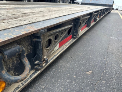 Flatbed trailer edge showing reinforced side rails, cutouts, and reflective red-and-white markings beneath a wooden deck. Visible are circular cutouts and lashing rings along the trailer side.