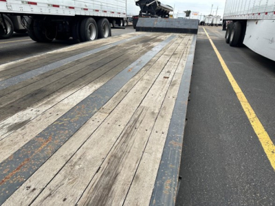Flatbed trailer bed with wooden planks and metal side rails, viewed from the rear with other trailers parked on both sides.