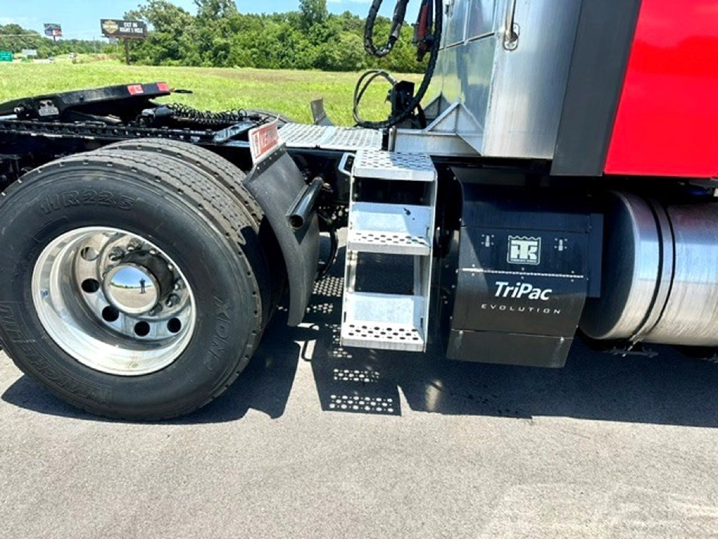 Truck wheel and step access near the cab of a 2022 Kenworth T680, showing a chrome fuel tank area, perforated metal steps, and a black TriPac Evolution battery/AC unit.