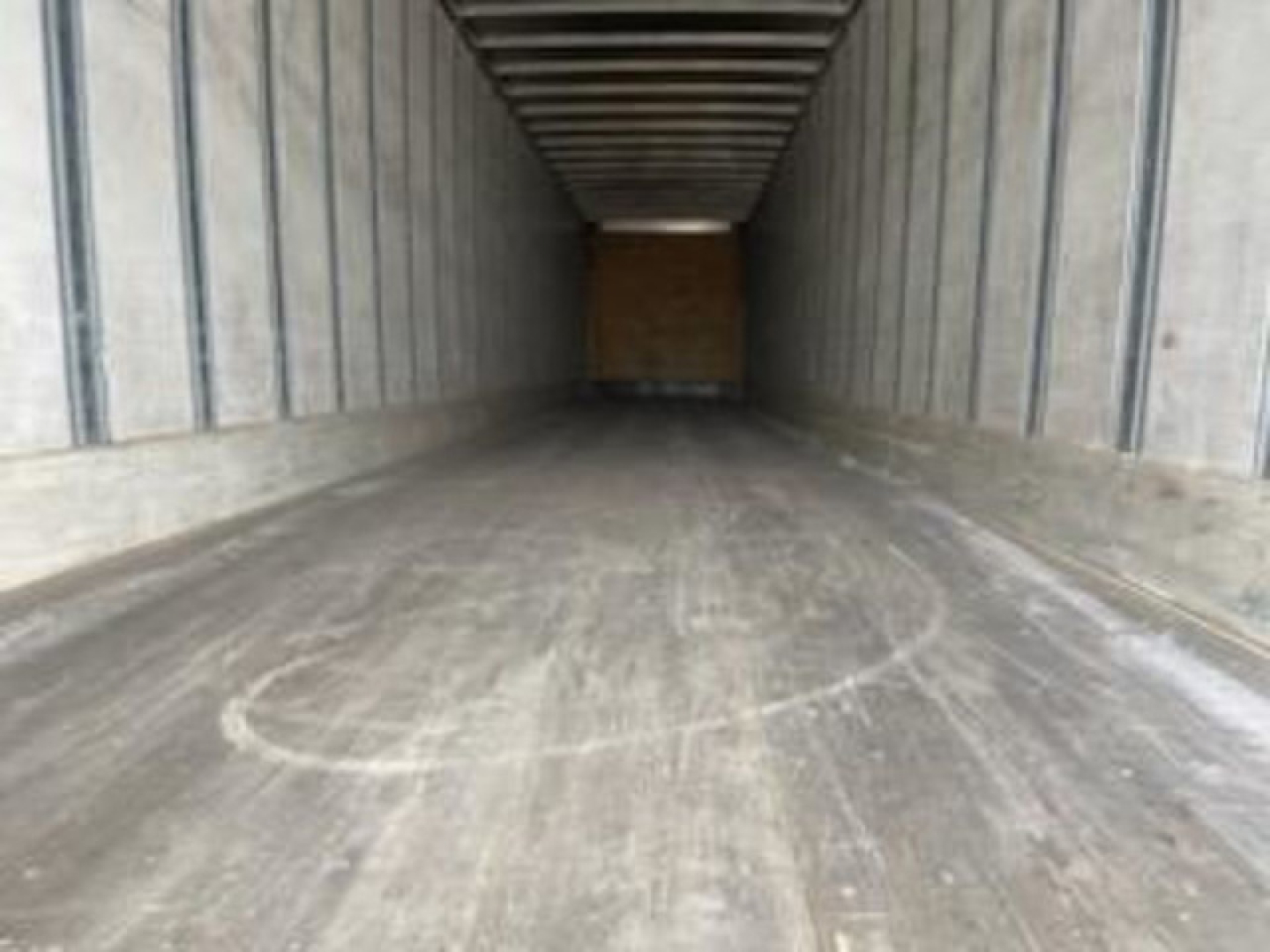 Inside of a long empty trailer with wooden front wall; concrete floor with wheel marks and circular scuff near foreground.