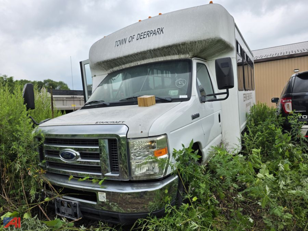 Fleet bus front view showing Ford E350 chassis with wheelchair bus conversion, front grille with Ford emblem, headlights, and overgrown vegetation around vehicle; text on top of bus reads 'TOWN OF DEERPARK'