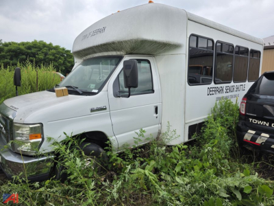 White bus with 'DEERPARK SERVICE' lettering on side, wheelchair-accessible bus, front cab with hood and large windshield, overgrown vegetation around wheel wells.