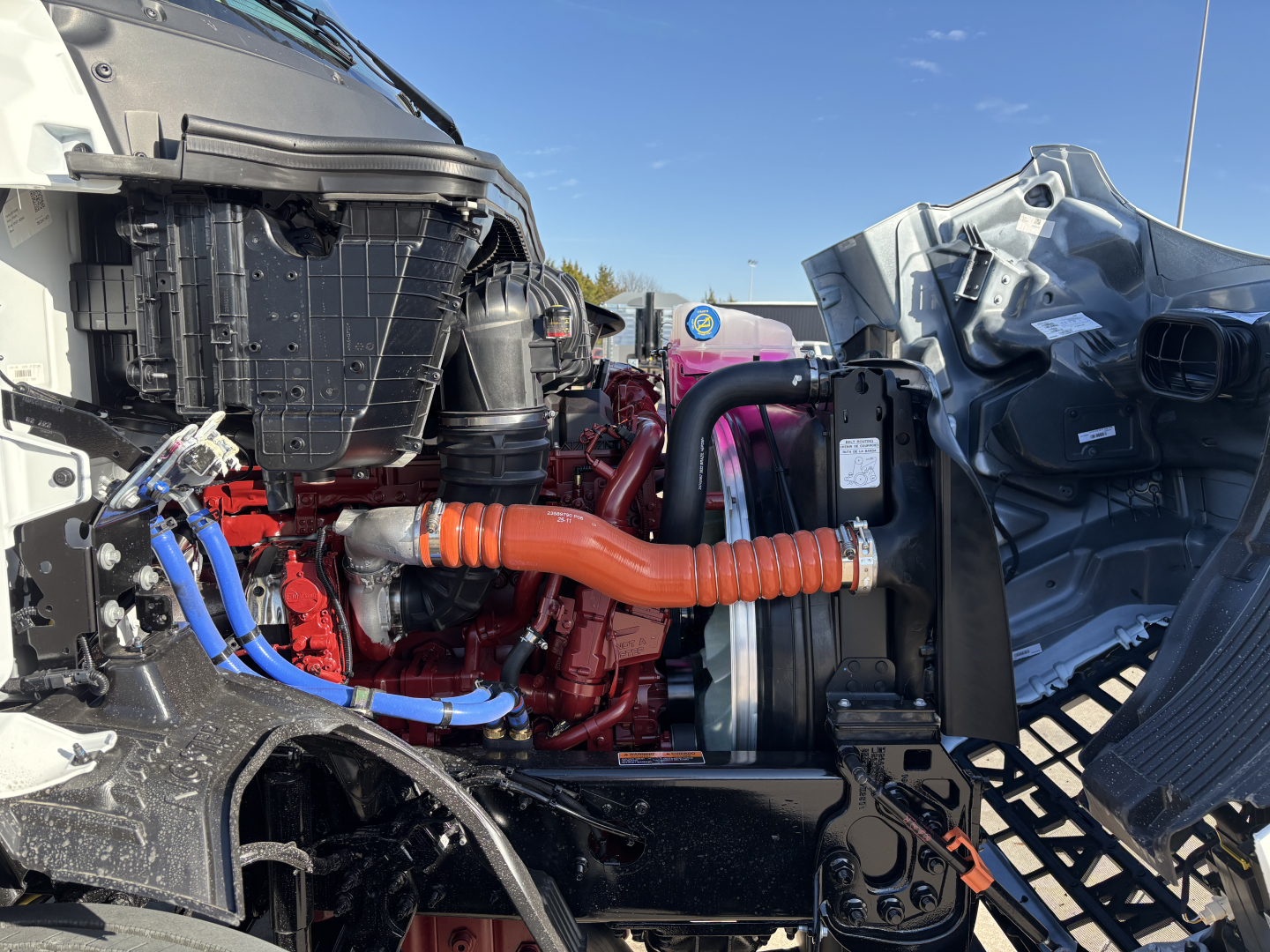 Trucked engine bay of a 2027 Mack AN64T showing red engine, orange coolant hose, blue lines, radiator, and surrounding frame with exposed components.
