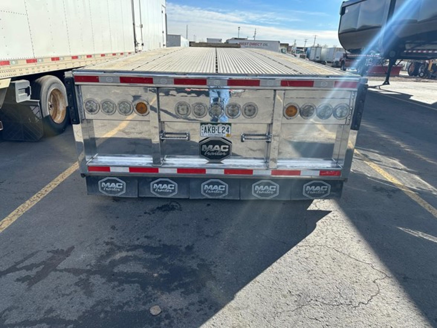 Flatbed trailer rear view with polished aluminum surface and MAC branding; multiple circular taillights, reflectors, and license plate visible.