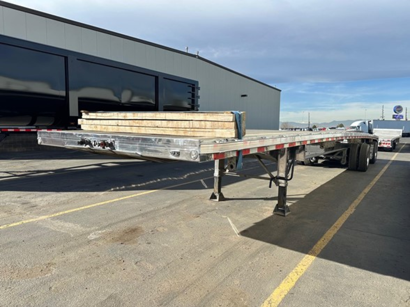 Flatbed trailer with polished aluminum finish showing extended bed and support legs, load of lumber on top, warehouse building in background.