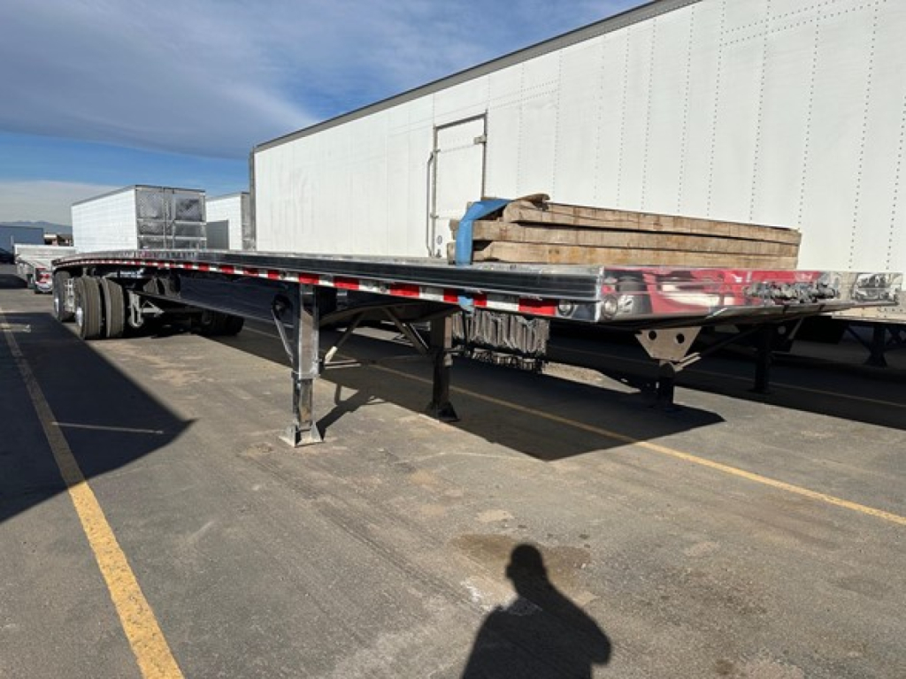 Flatbed trailer with polished aluminum deck and undercarriage, extended length, side rails, and landing gear visible.
