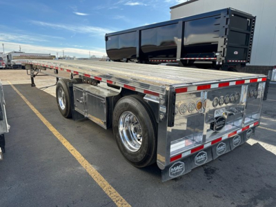 Mac polished all-aluminum flatbed trailer with dual rear axles and chrome wheels, polished metal deck, rear storage/tool boxes, and rear bumper/lighting assembly.