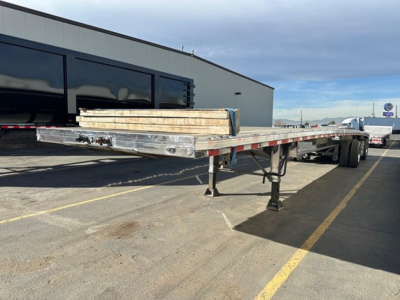 Flatbed trailer with polished aluminum finish showing extended bed and support legs, load of lumber on top, warehouse building in background.
