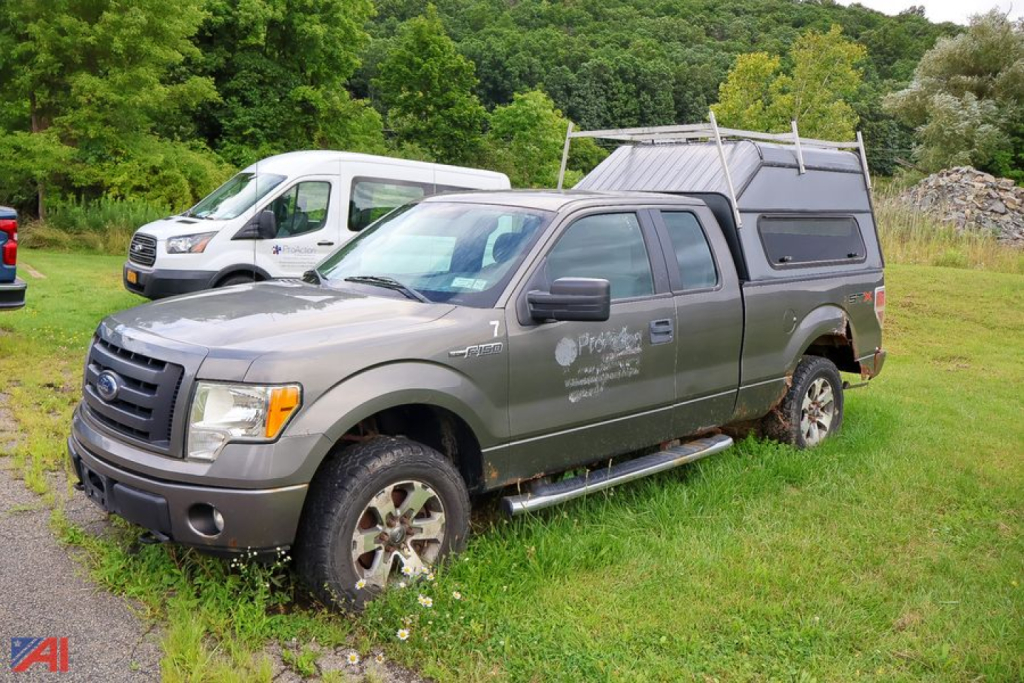 Ford F-150 crew cab pickup with cap, gray exterior, four-wheel drive, off-road tires, side step bars, in a grassy lot.