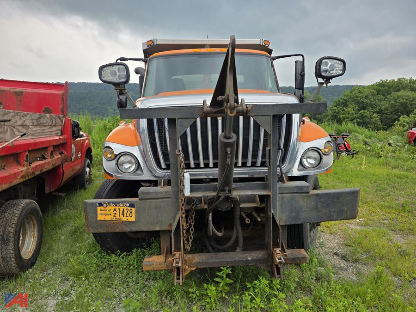Dump truck front view with heavy plow/frame attachment, International WorkStar 7500, orange cab, yellow license plate, in grassy lot.