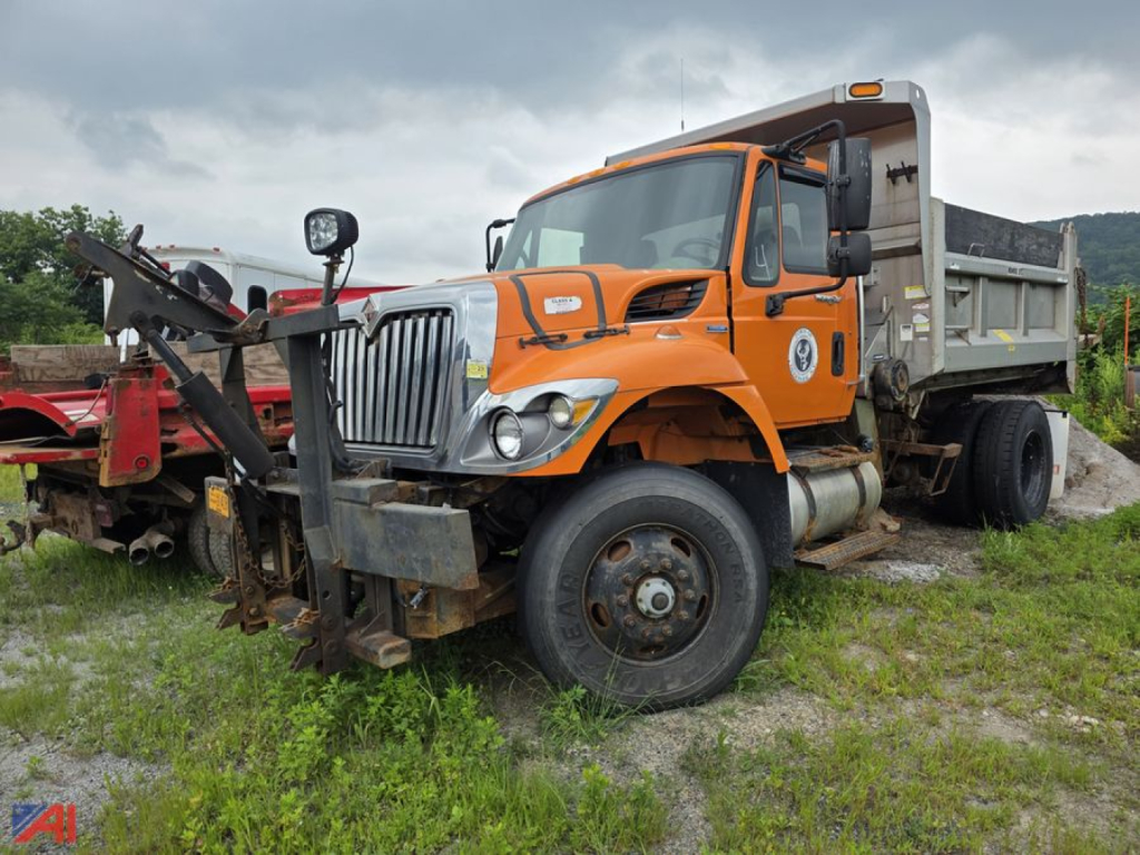 Orange International WorkStar 7500 dump truck viewed from front-left, with heavy front bumper and attached digging/loader equipment, dual rear wheels and open cargo bed