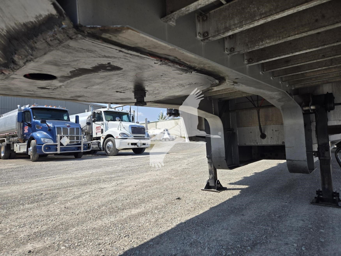 Underneath view of a 2019 Reitnouer Dropmiser trailer showing undercarriage with support legs and frame; background shows two trucks in a lot.