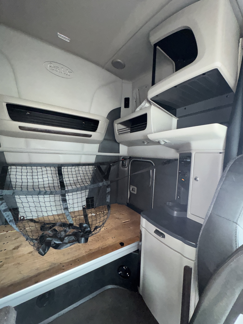 Peterbilt 579 cabin interior showing bunk area and overhead storage, wood floor base, driver seat, netting under bunk, dash area, and storage compartments.