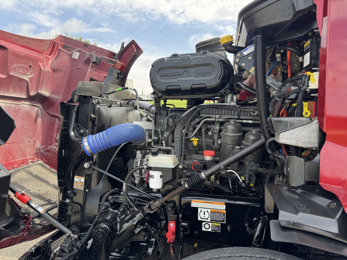 Peterbilt 579 engine bay with visible air intake, coolant reservoir, hoses, and wiring; front left fender open showing engine components and battery area.