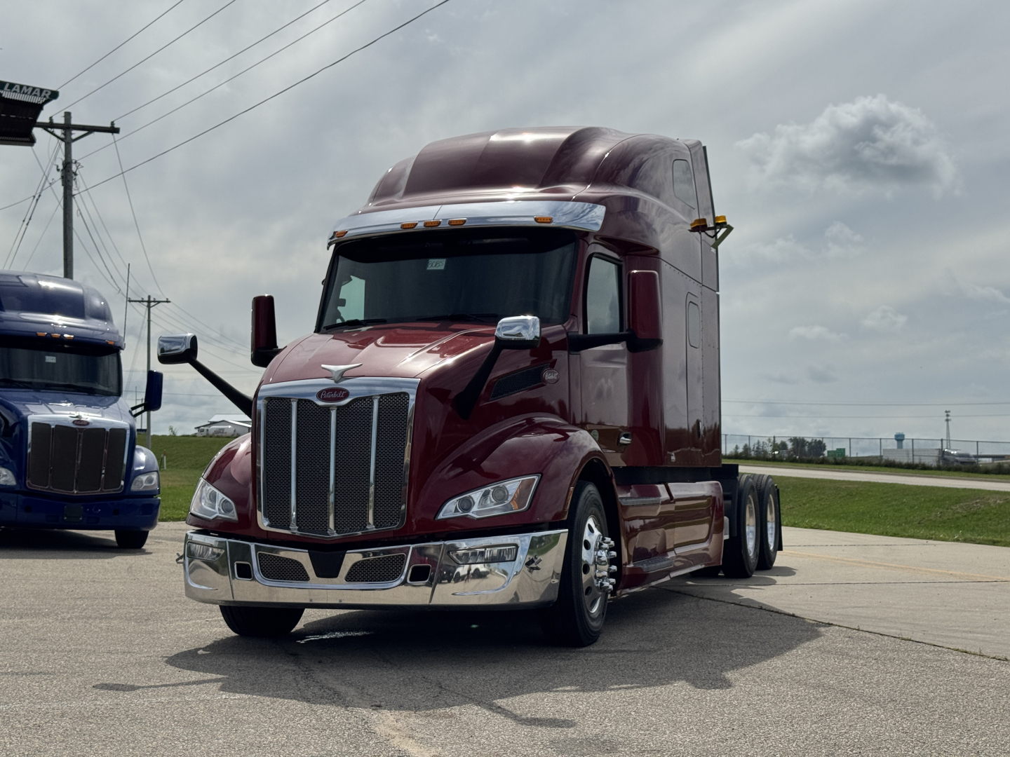 Peterbilt 579 truck in a parking lot with maroon cab, chrome grille and mirrors, black tires, blue truck in background. visible text not legible.