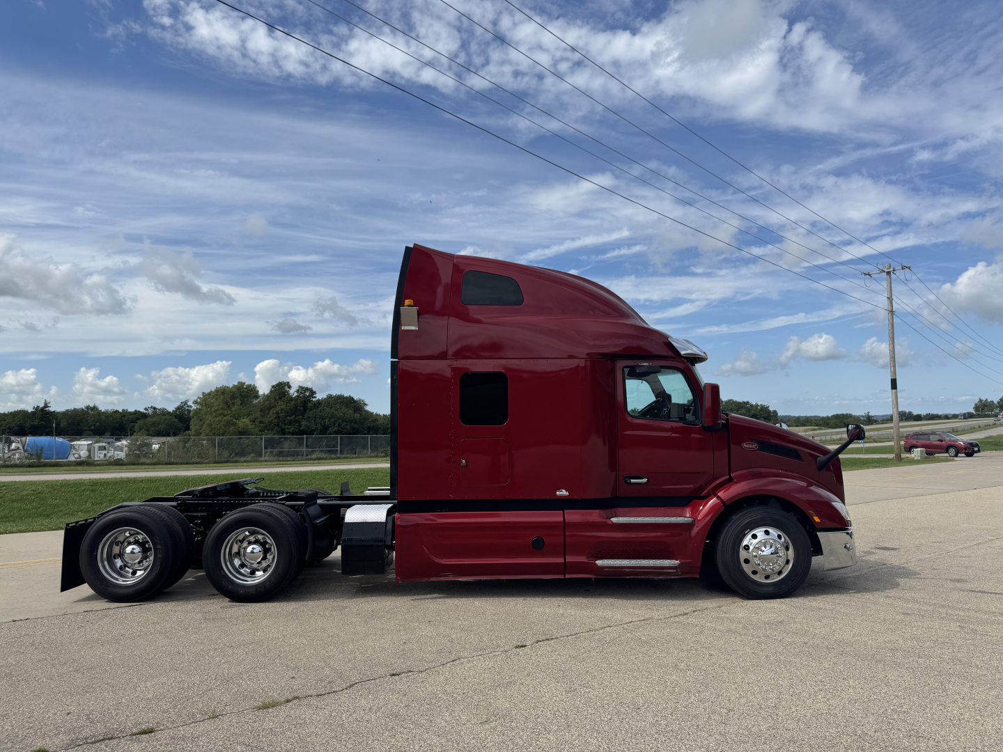 Truck side view of a red Peterbilt 579 tractor, sleeper cab, five tires on the rear, clean and polished, with blue sky and clouds.