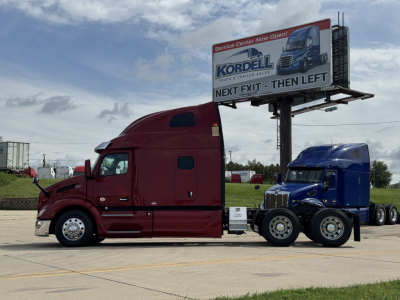 Peterbilt 579 semi truck in maroon with a blue tractor in the background, billboard trailer, highway with service center sign visible.