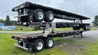 Trailer stack showing a Manac steel flatbed trailer with dual axles and black frame; visible wheels, reflective safety stripes, and mounting hardware.