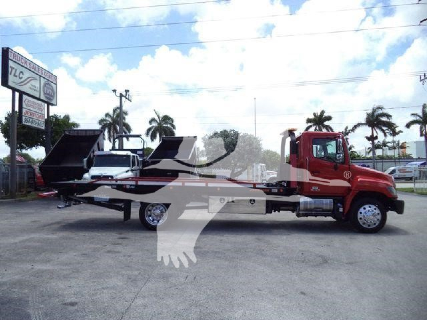 Truck: 2020 Hino 268A rollback w/ flatbed and tilting mechanism, red cab, black flatbed, side view. Text on sign in background reads TLC Truck Sales. Palm trees and clear sky.