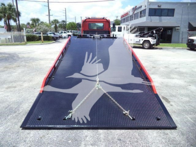 Flatbed truck bed of a 2020 Hino 268A rollback with a large dove graphic on the deck, rear view, black textured surface, chains securing the bed, red cab in the background.