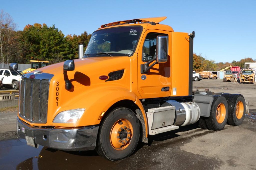 Peterbilt 579 daycab truck in orange, front-left view, with dual rear axles, side fuel tanks, black grille, and model badges on hood.