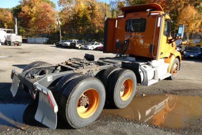 Peterbilt 579 day cab tractor rear view showing chassis, dual rear axles, and white hitch, orange cab, visible mud puddle in lot
